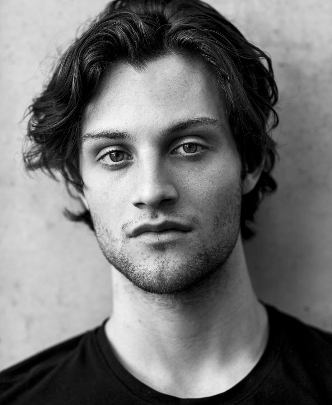 A black and white portrait of a young man with dark wavy hair in front of a concrete wall